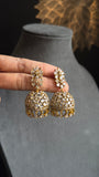 Gold and crystal earrings held by a hand on a gray surface with a blurred decorative background.