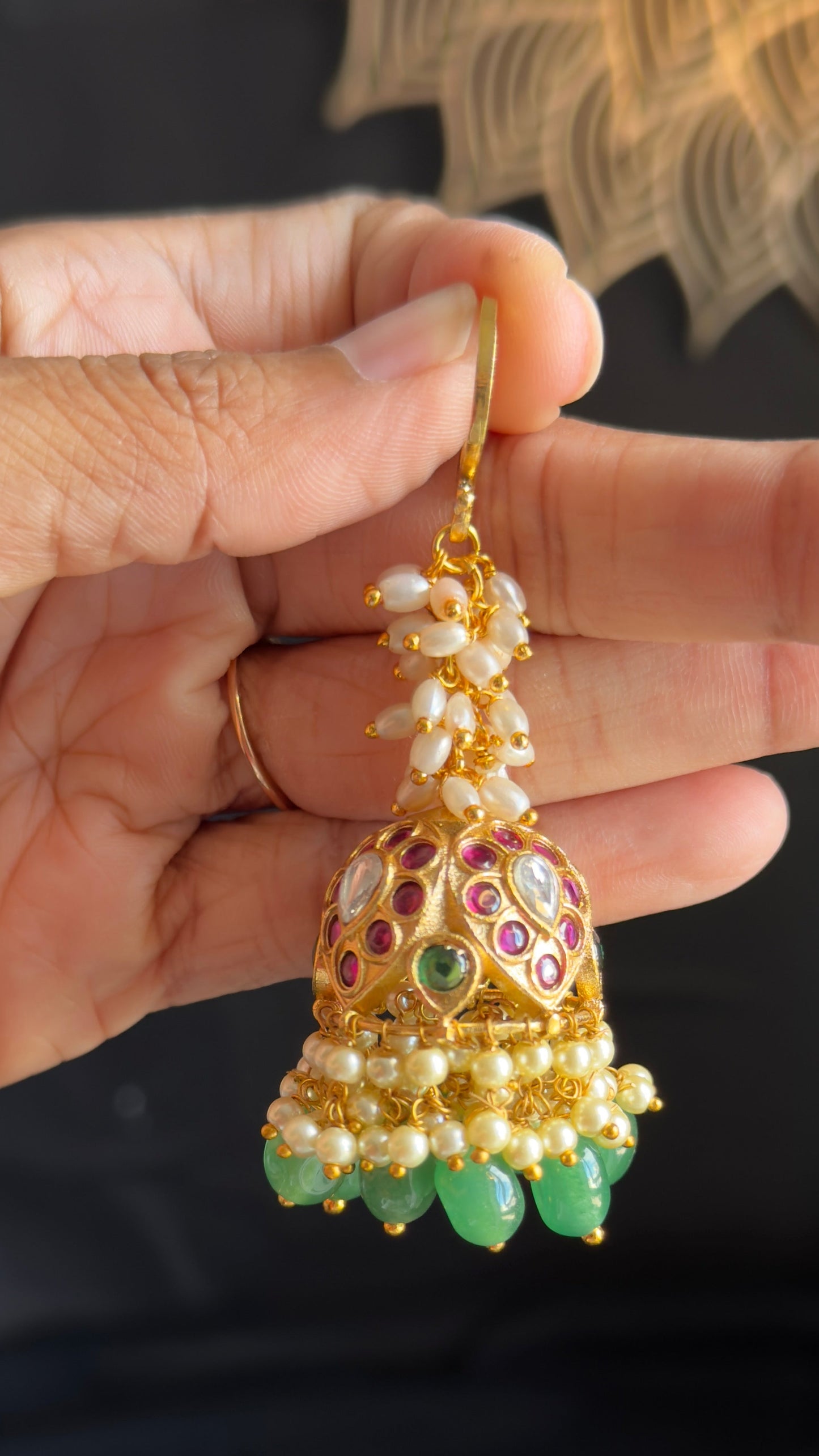 A hand holding a gold-toned traditional Indian jhumka earring decorated with white pearls, green stones, and red and purple enamel details.