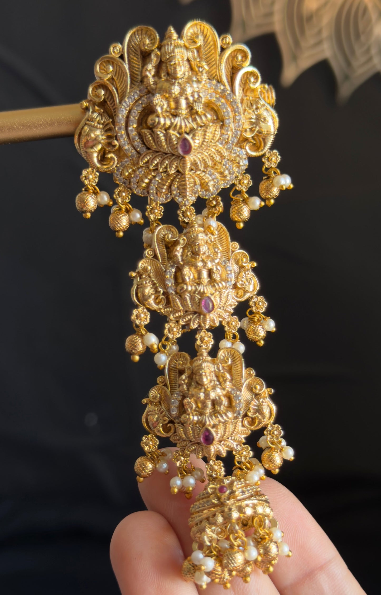 An ornate gold-toned traditional Indian jewelry piece with intricate filigree work, small bead drops, and dark red stone accents, held in a person's hand against a dark background.