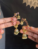 A pair of gold-plated Indian jhumka earrings with pink and green enamel, peacock motifs, and white pearl bead borders, held up by a person's hands against a black shirt.