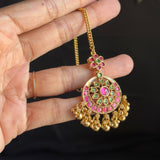 A hand holding a gold-toned traditional Indian jhumka necklace with pink and green stone embellishments against a dark background.