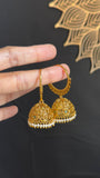 A pair of gold-toned filigree jhumka earrings with small white bead accents and subtle blue-green stone details, held by a hand against a dark background with a decorative gold motif.