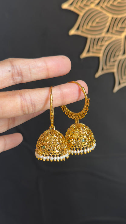 A pair of gold-toned filigree jhumka earrings with small white bead accents and subtle blue-green stone details, held by a hand against a dark background with a decorative gold motif.