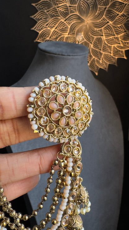 A close-up of a hand holding Panoplylondon's intricately designed 2-in-1 Detachable Antique Gold Tone Earrings with Chain, Polkis, and Faux Pearls. The circular earrings in an antique gold tone feature pearl and gemstone accents with tassel-like chains hanging below. In the background, there's a black mannequin and wall decor resembling a floral pattern.