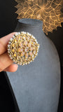 A person holds a pair of ornate, detachable earrings with intricate gold details, faux pearl accents, and polkis. The antique gold tone earrings from Panoplylondon are showcased against a gray mannequin bust in a dimly lit setting, with decorative floral patterns in the background.