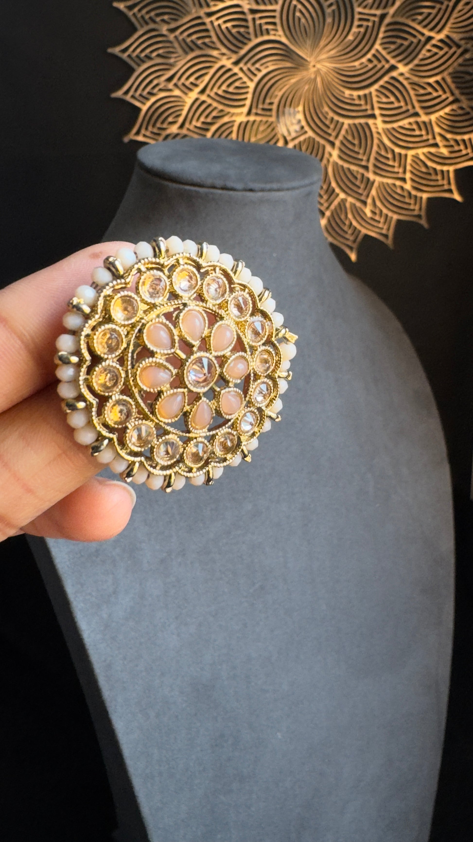 A person holds a pair of ornate, detachable earrings with intricate gold details, faux pearl accents, and polkis. The antique gold tone earrings from Panoplylondon are showcased against a gray mannequin bust in a dimly lit setting, with decorative floral patterns in the background.