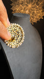 A person holds a pair of intricately designed 2-in-1 Detachable Antique Gold Tone Earrings, featuring chains, polkis, and faux pearls from Panoplylondon, in front of a black mannequin stand. The background showcases an elegant, illuminated floral mandala design that evokes the rich heritage found in Indian and Pakistani outfits.