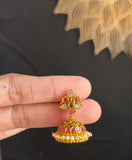 A hand holding a small gold-toned traditional Indian jhumka earring decorated with red, green, and white stones, set against a dark black fabric background.