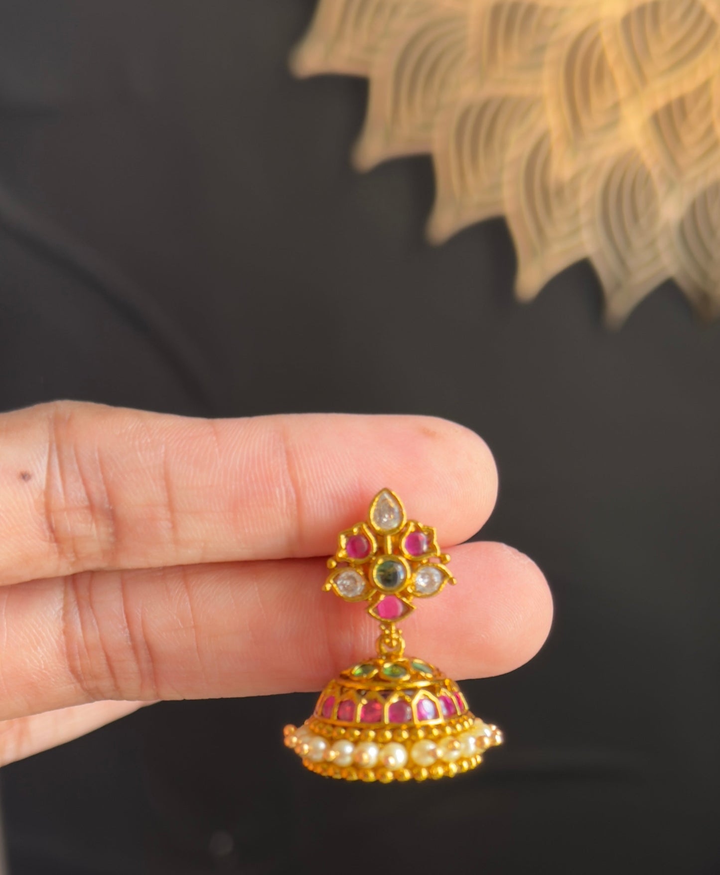 A hand holding a small gold-toned traditional Indian jhumka earring decorated with red, green, and white stones, set against a dark black fabric background.