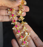 A pair of gold-toned traditional Indian earrings with pink stones and white pearl-like beads, held in a person's hand against a dark background.