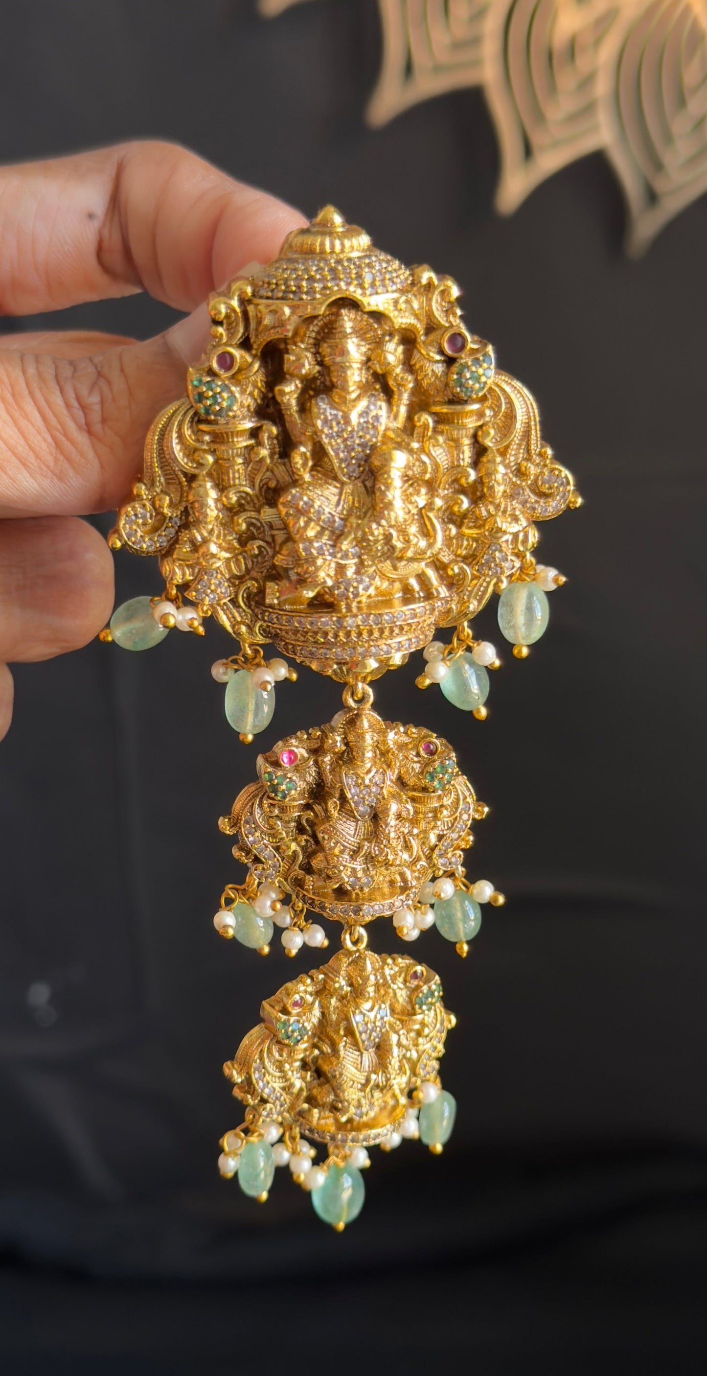 A hand holding a large, ornate gold-toned Ganesha pendant or brooch with dangling green bead accents, set against a dark fabric background.
