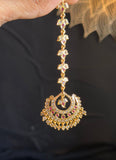 A gold-toned traditional Indian maang tikka with small pink and green stone accents, held by a hand against a dark background.