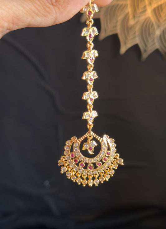 A gold-toned traditional Indian maang tikka with small pink and green stone accents, held by a hand against a dark background.