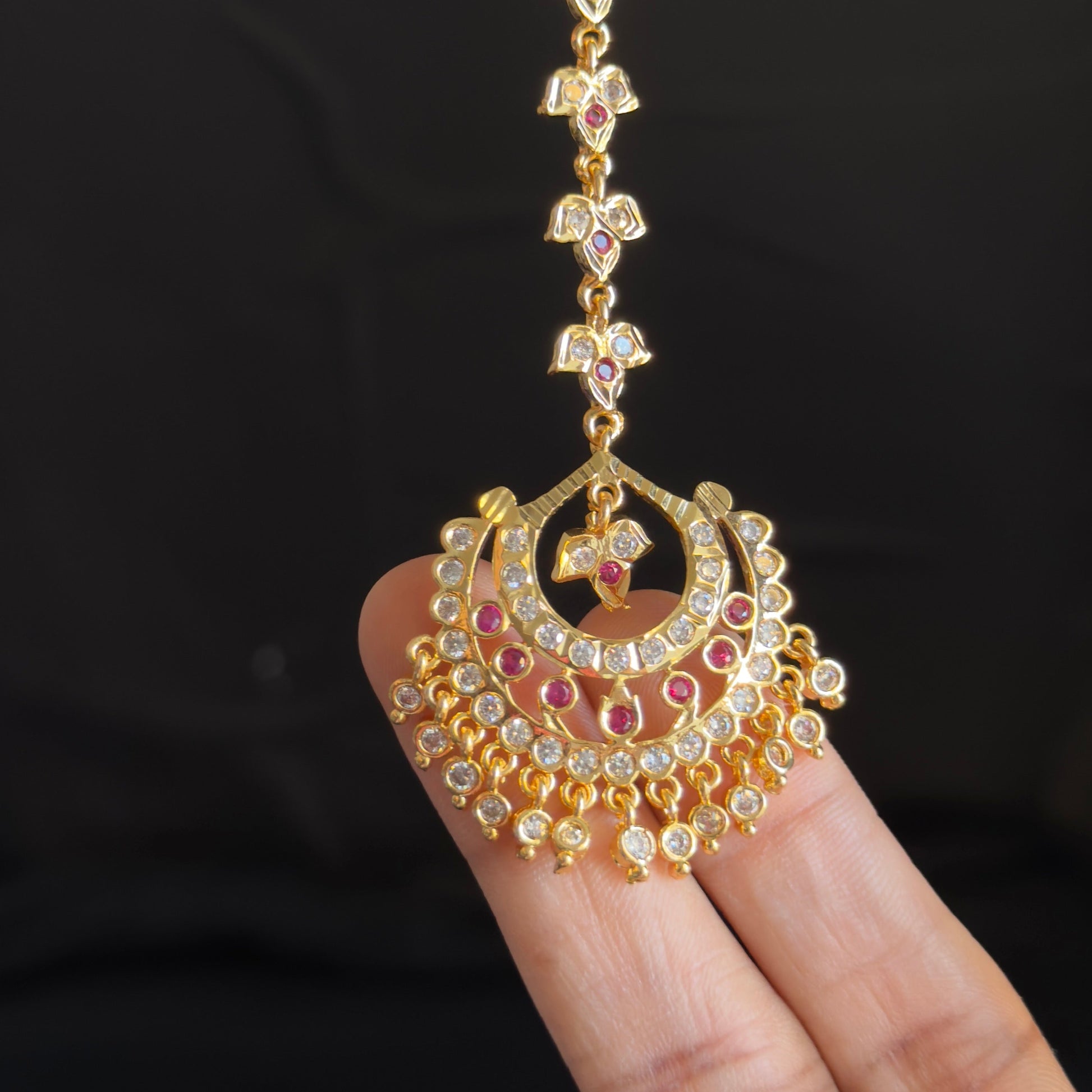 A gold-toned traditional Indian jhumka earring with red and white stones, held by a person's hand against a dark background.