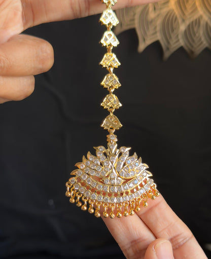 A close-up of a gold-toned traditional Indian jhumka earring being held by hands against a dark background.