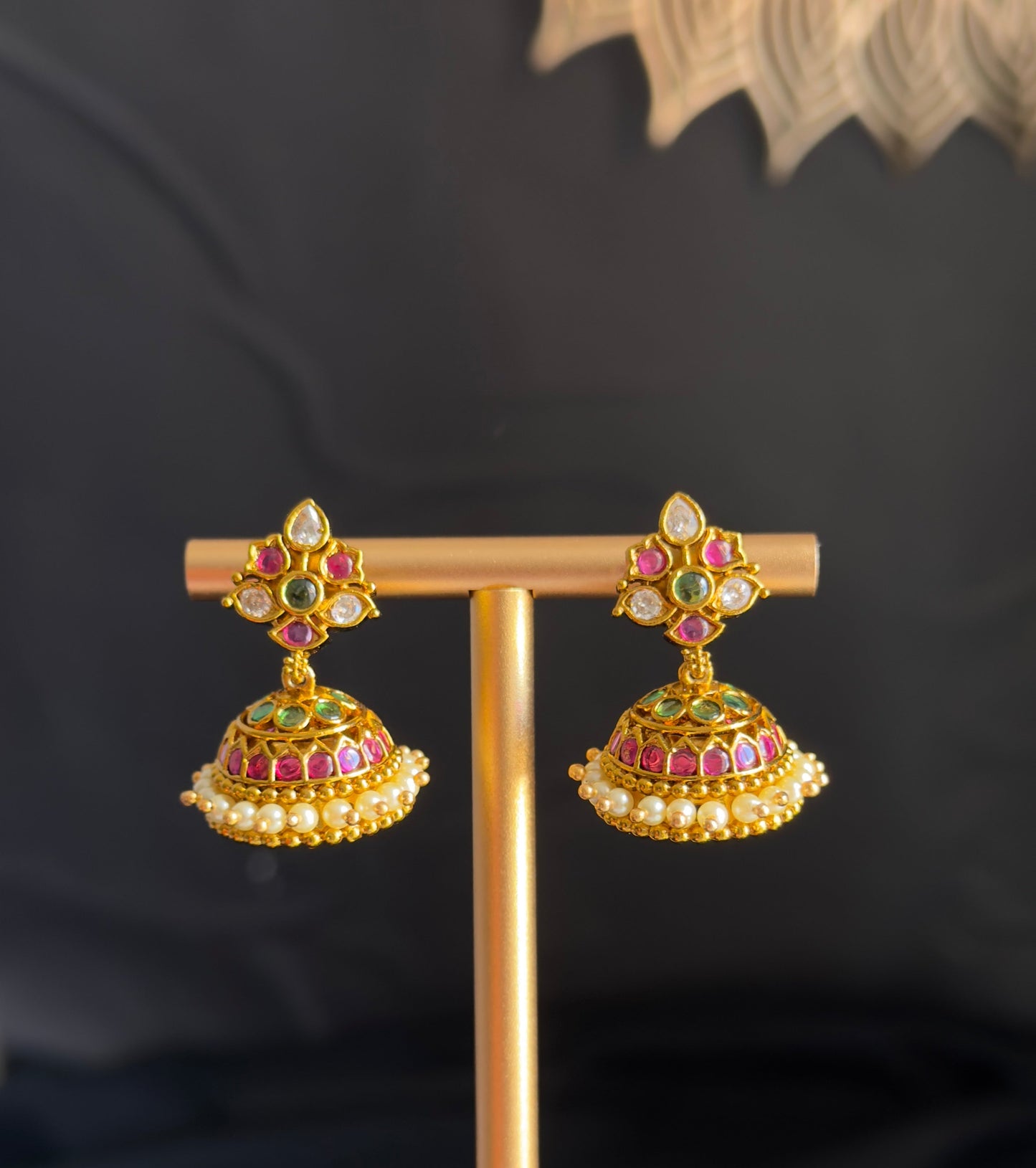 A pair of gold-toned traditional Indian jhumka earrings with red, green, and white stone embellishments, displayed on a gold stand against a dark background.