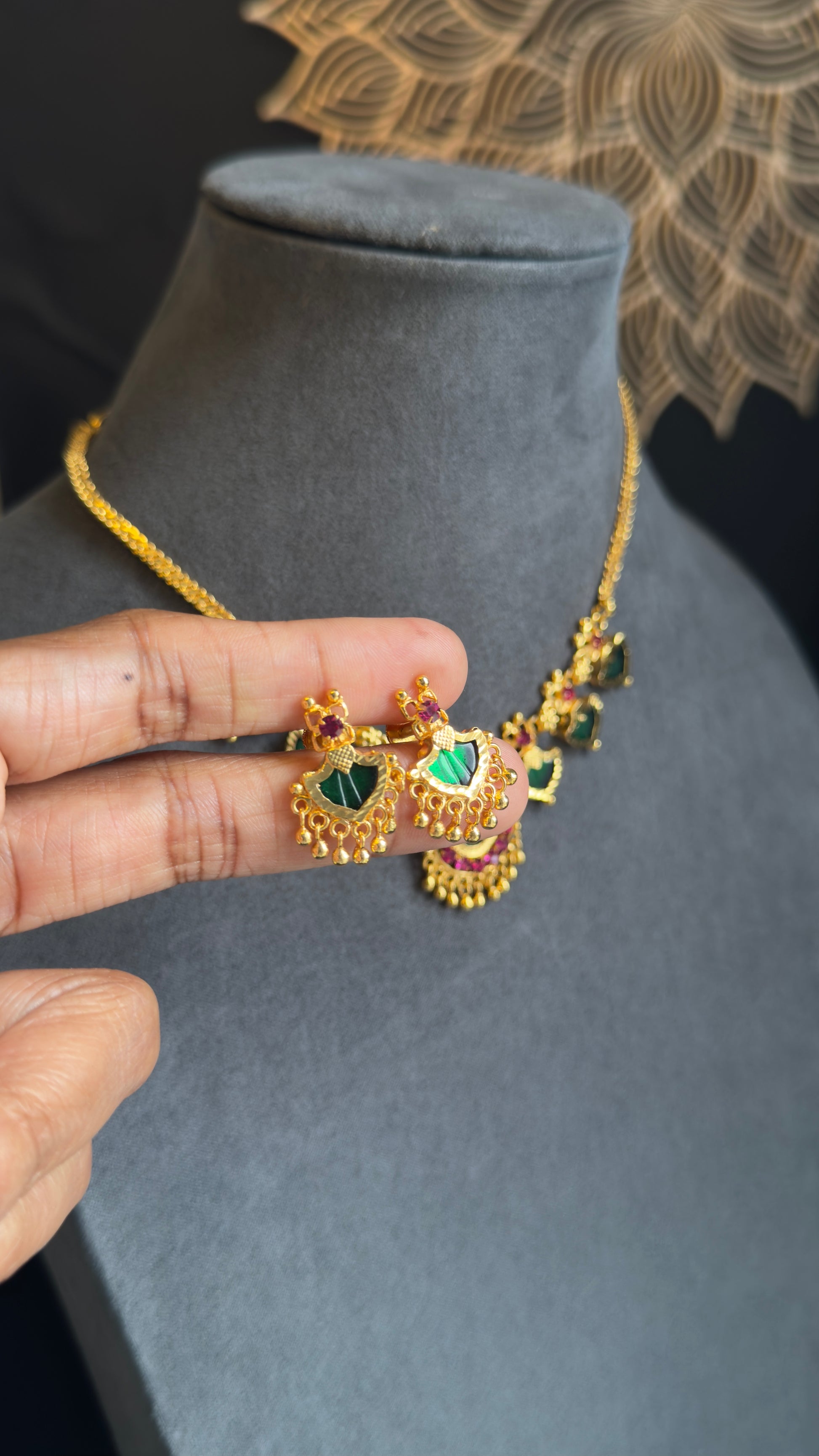 Gold necklace with green gemstones held by a hand on a gray mannequin bust.