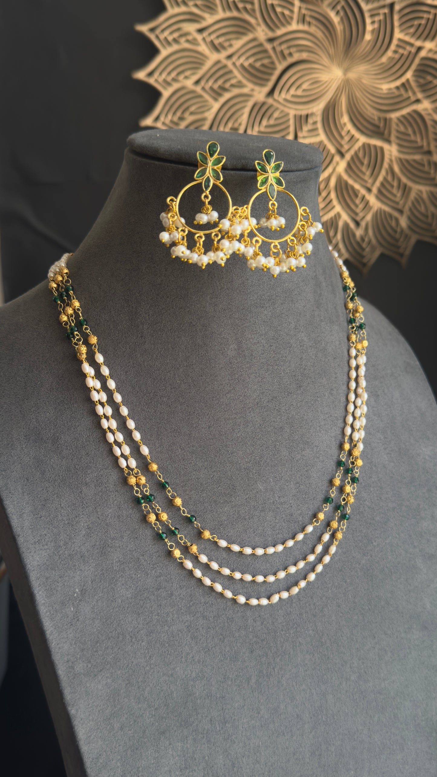 A three-layered pearl and bead necklace set with matching gold-toned earrings, displayed on a gray jewelry bust against a dark background with a decorative mandala motif.