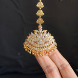 A gold-toned traditional Indian jhumka necklace with a detailed paisley pendant, held by a person's fingers against a dark background.