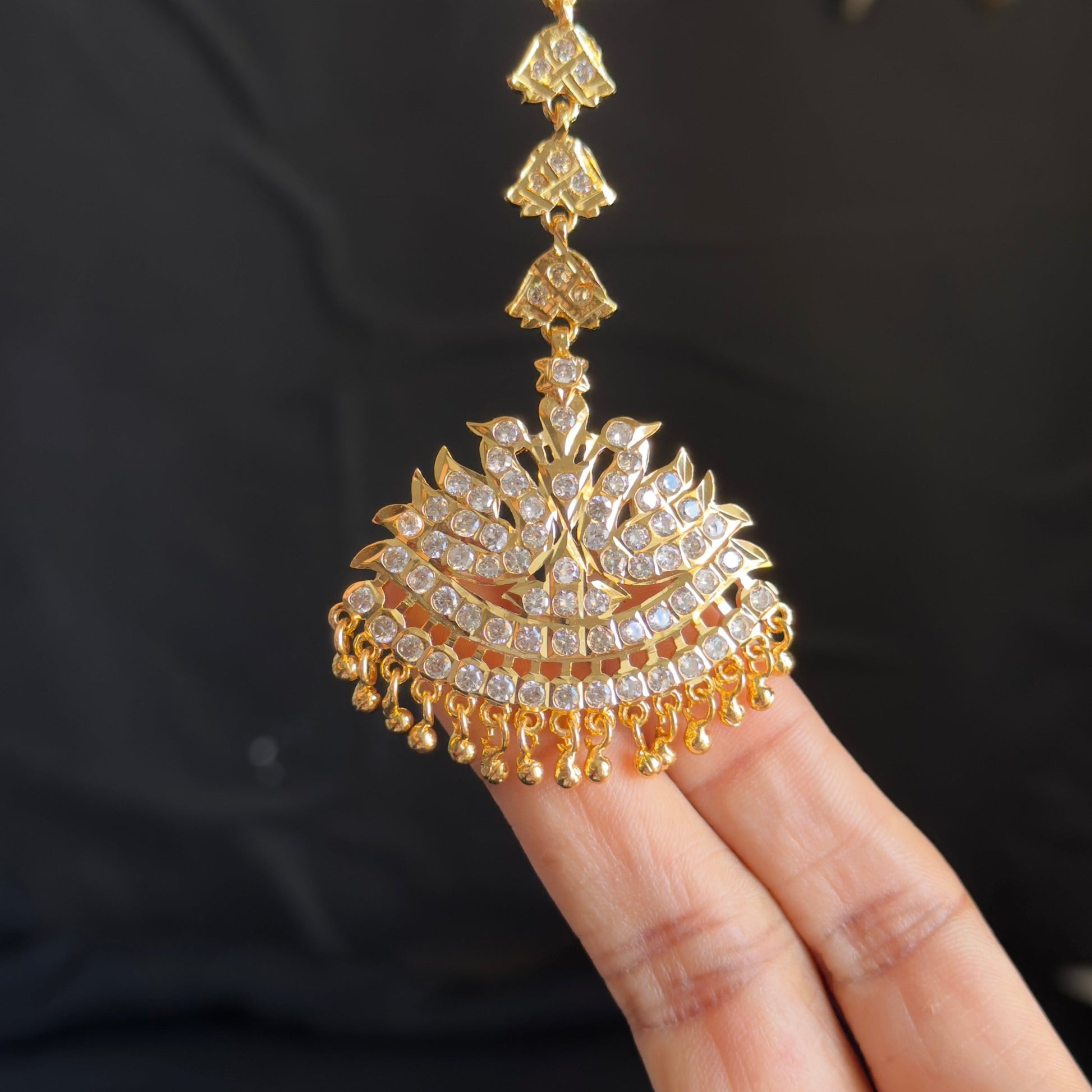 A gold-toned traditional Indian jhumka necklace with a detailed paisley pendant, held by a person's fingers against a dark background.