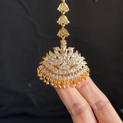 A gold-toned traditional Indian jhumka necklace with a detailed paisley pendant, held by a person's fingers against a dark background.