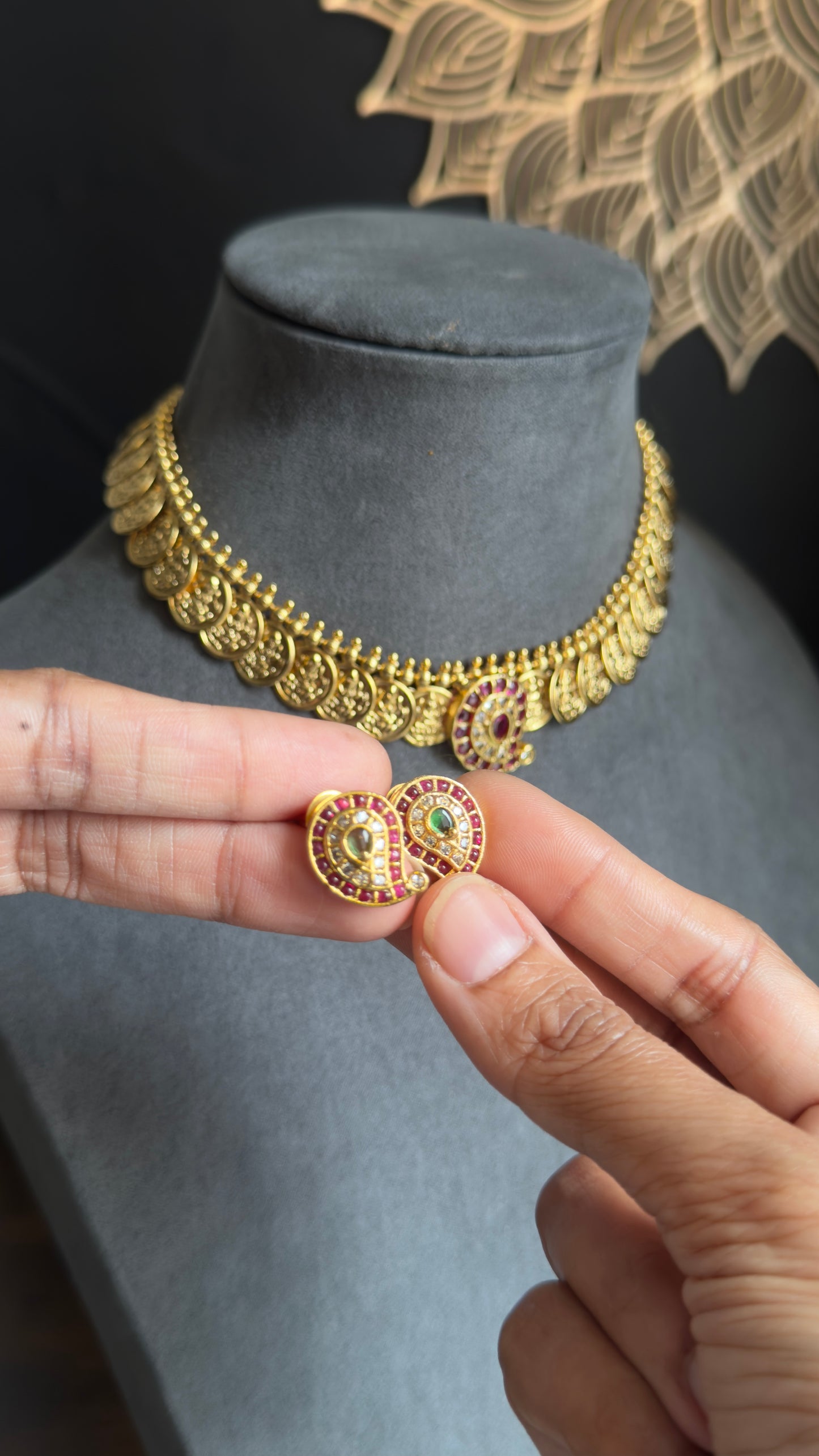 Gold necklace on a mannequin bust with a hand holding a pair of gold earrings with gemstones.