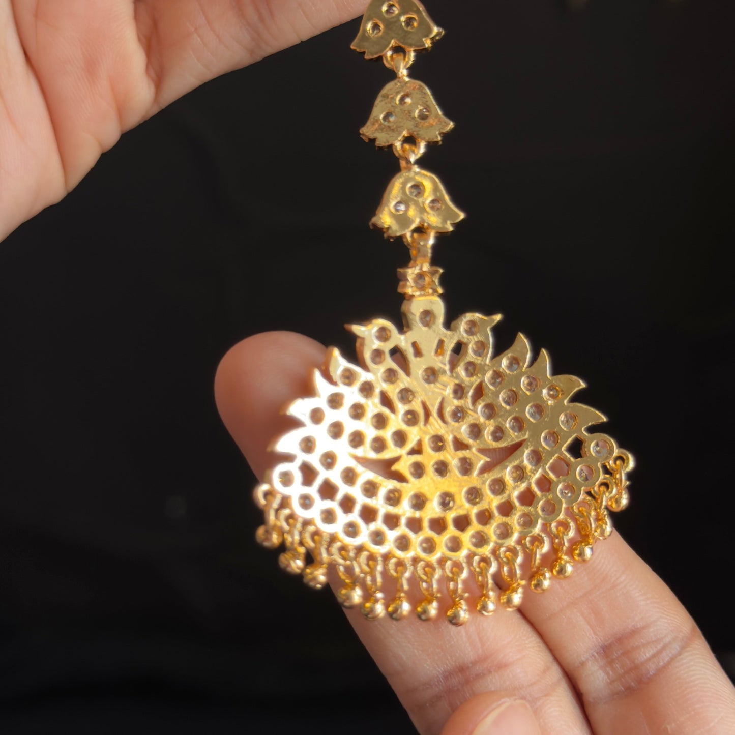 A close-up of a hand holding a gold-toned traditional Indian Jhumka earring with a detailed filigree design and dangling bead chain against a dark background.