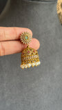 A hand holding a gold-toned traditional Indian jhumka earring with a green stone center, rhinestone-encrusted circular top, and dangling bell-shaped charm with white pearl beads at the bottom, set against a gray jewelry display bust.