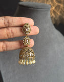 A hand holds a gold-toned traditional Indian jhumka earring with white stone embellishments and dangling pearl-like beads, positioned in front of a gray jewelry display bust.