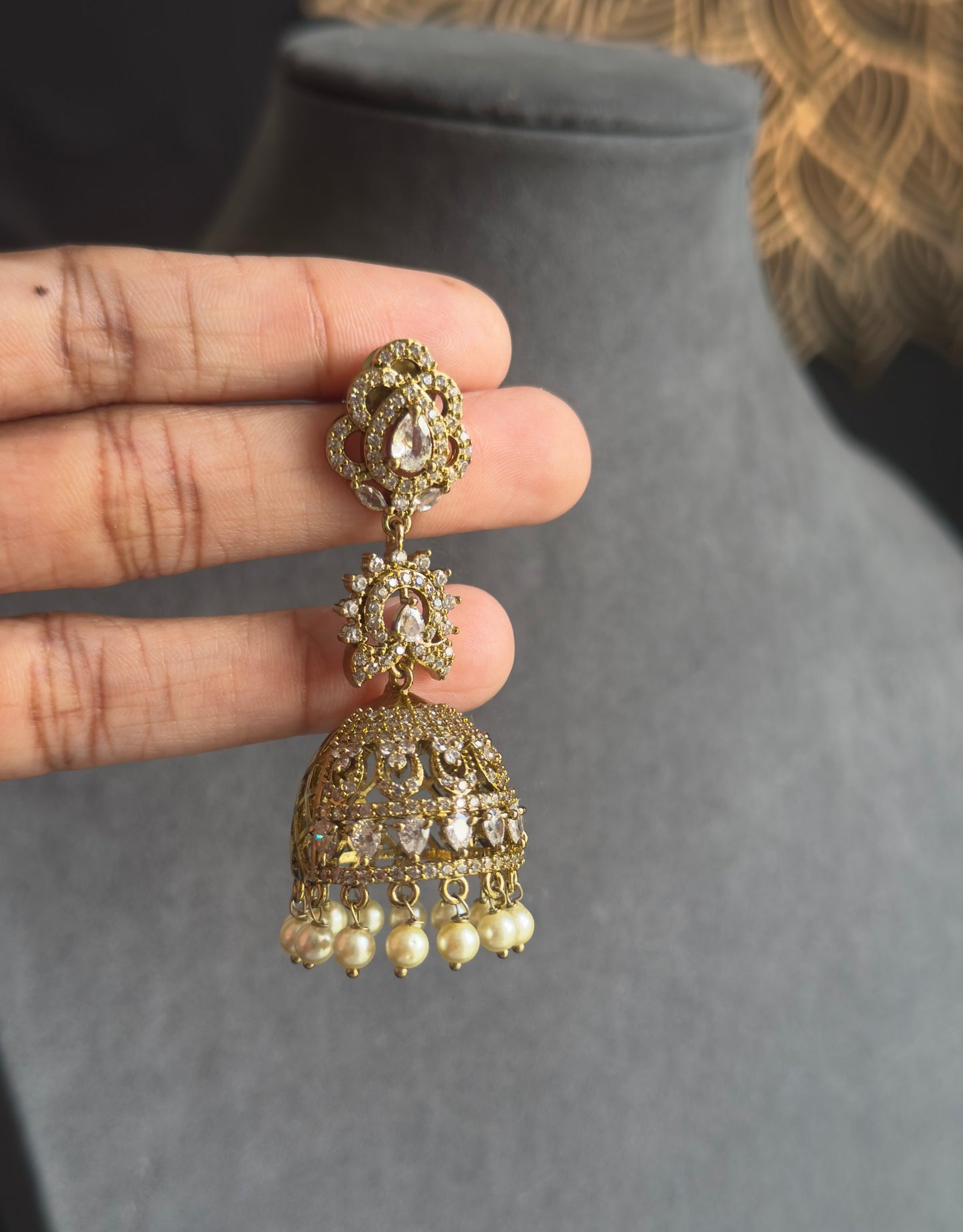 A hand holds a gold-toned traditional Indian jhumka earring with white stone embellishments and dangling pearl-like beads, positioned in front of a gray jewelry display bust.