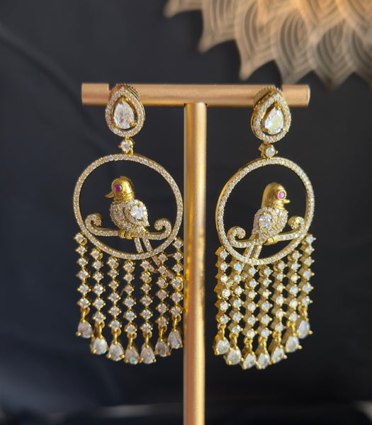 A pair of gold-toned traditional Indian chandbali earrings with bird motifs and dangling bead chains, displayed on a gold stand against a dark background.