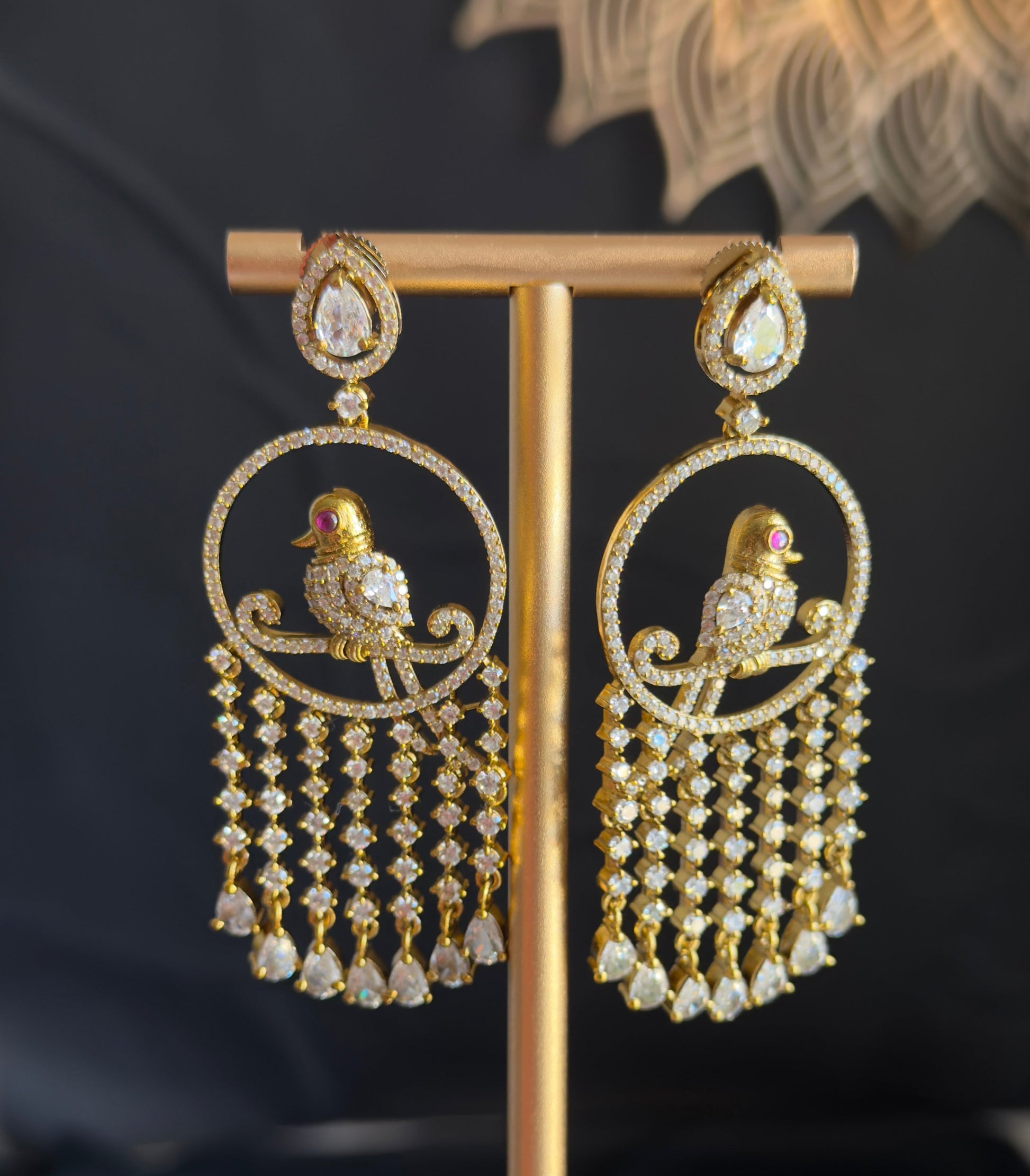 A pair of gold-toned traditional Indian chandbali earrings with bird motifs and dangling bead chains, displayed on a gold stand against a dark background.