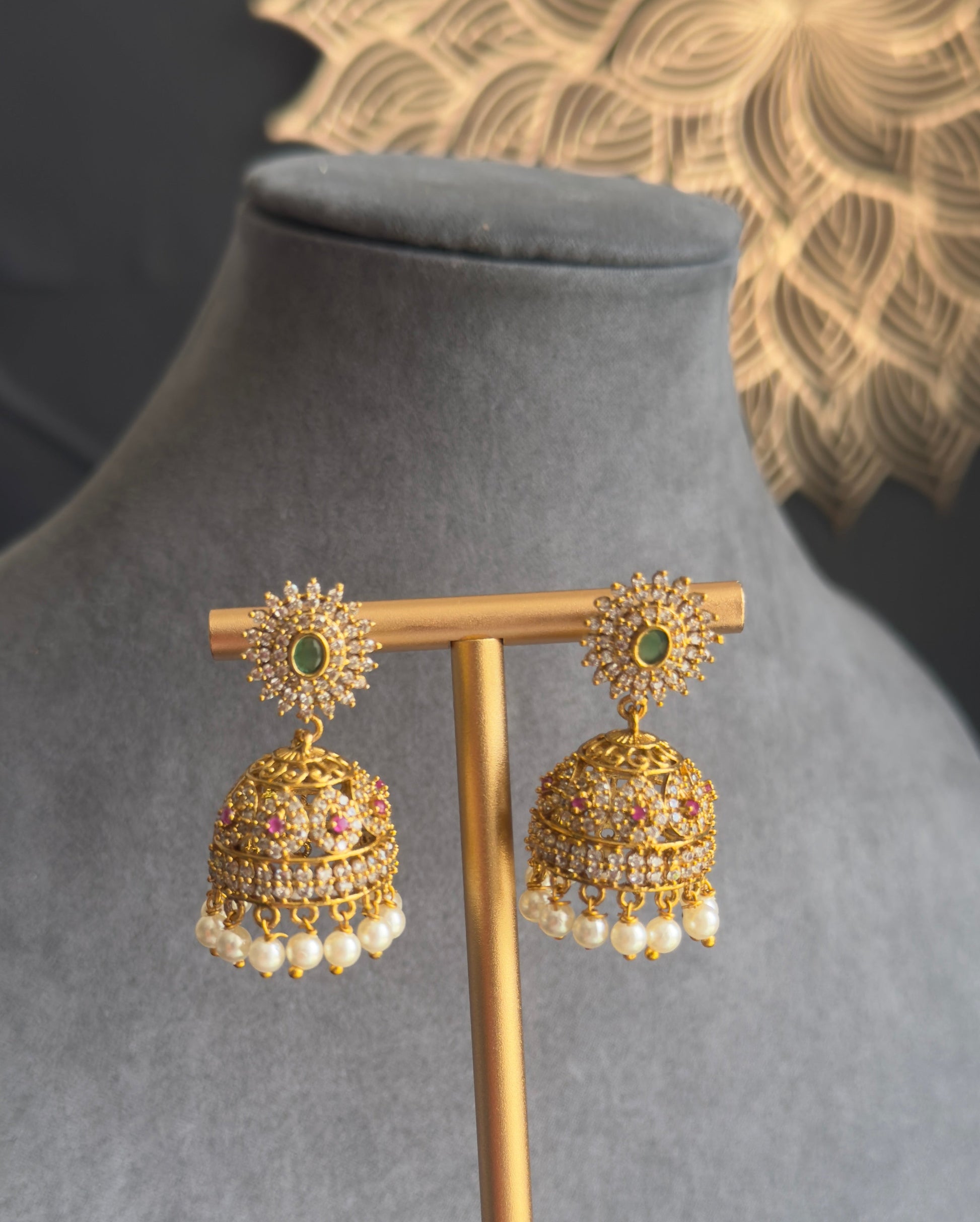 A pair of gold-toned jhumka earrings with green stone accents and dangling pearl-like beads, displayed on a gold stand against a gray velvet jewelry bust.
