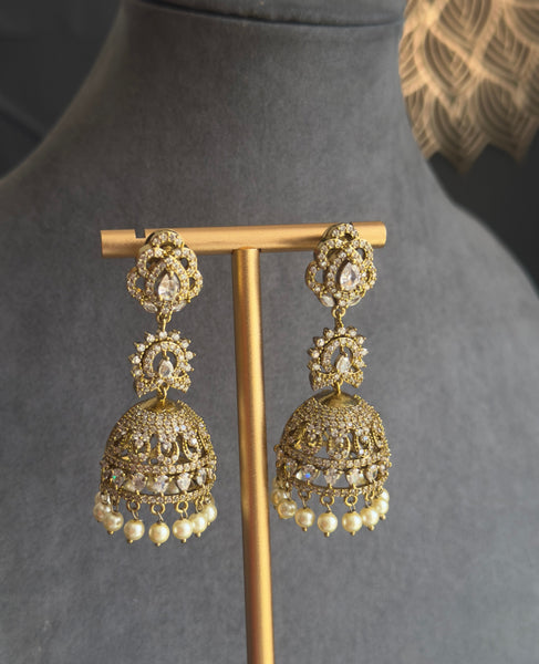 A pair of ornate gold-toned jhumka earrings displayed on a wooden stand against a dark gray velvet jewelry bust. The earrings feature intricate filigree detailing and small white bead drops at the bottom.