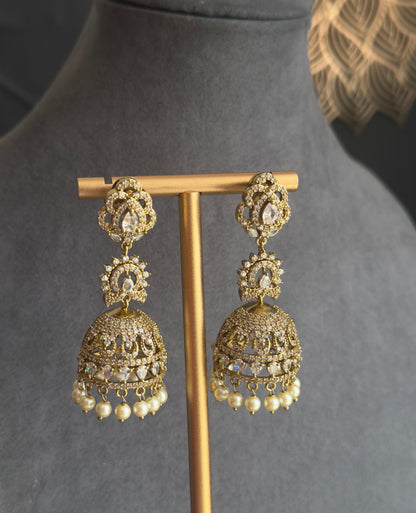 A pair of ornate gold-toned jhumka earrings displayed on a wooden stand against a dark gray velvet jewelry bust. The earrings feature intricate filigree detailing and small white bead drops at the bottom.