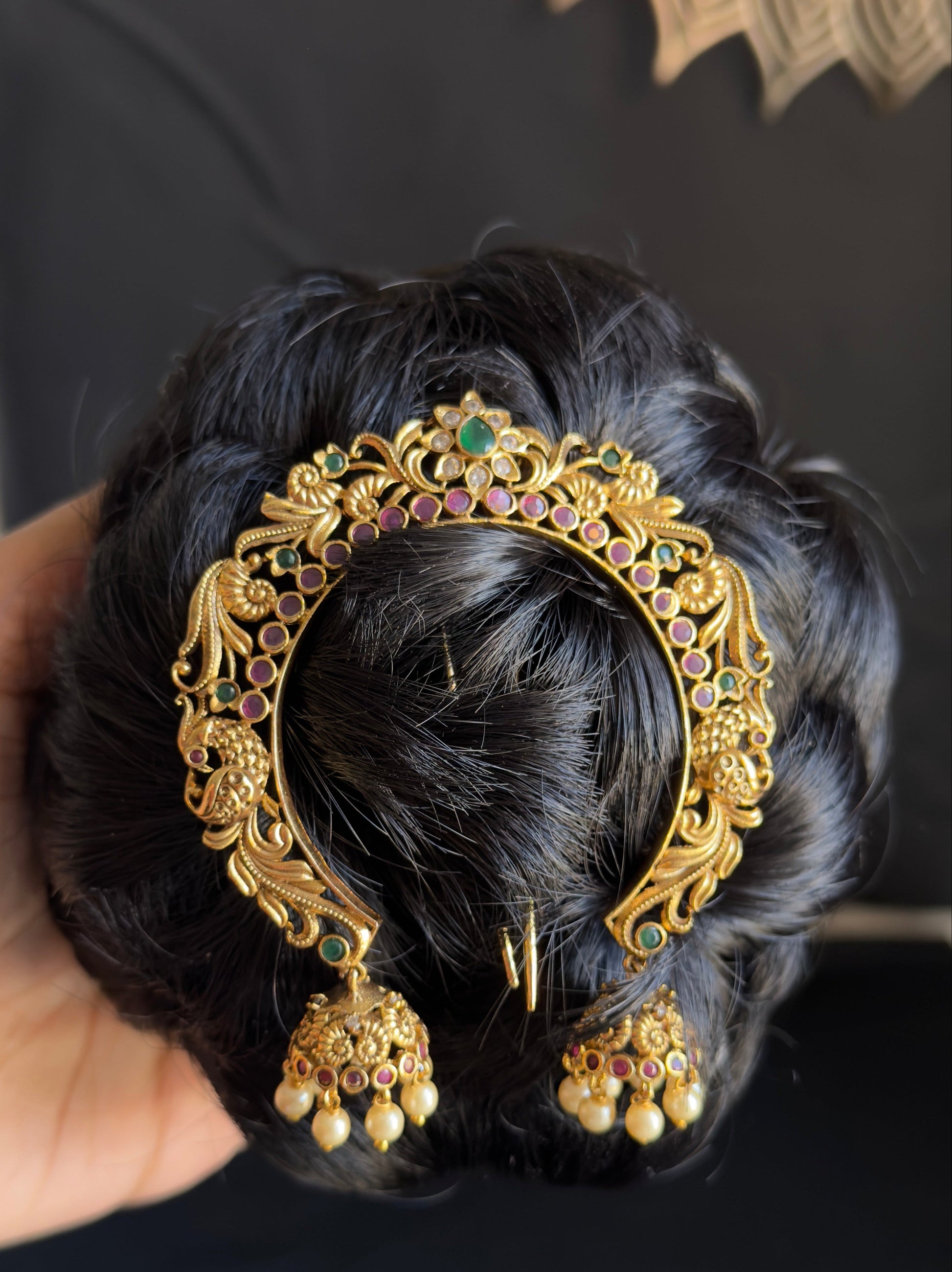 An ornate gold-toned traditional Indian hair accessory with filigree detailing, small green and pink stones, and dangling jhumka-style charms, shown pinned to a black hair bun held by a hand.