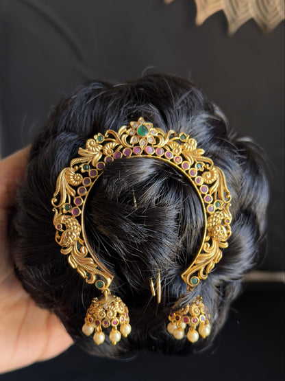 An ornate gold-toned traditional Indian hair accessory with filigree detailing, small green and pink stones, and dangling jhumka-style charms, shown pinned to a black hair bun held by a hand.