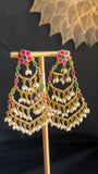 A pair of ornate gold-toned Indian jhumka earrings with white pearl bead drops and pink and green stone accents, displayed on a gold stand.