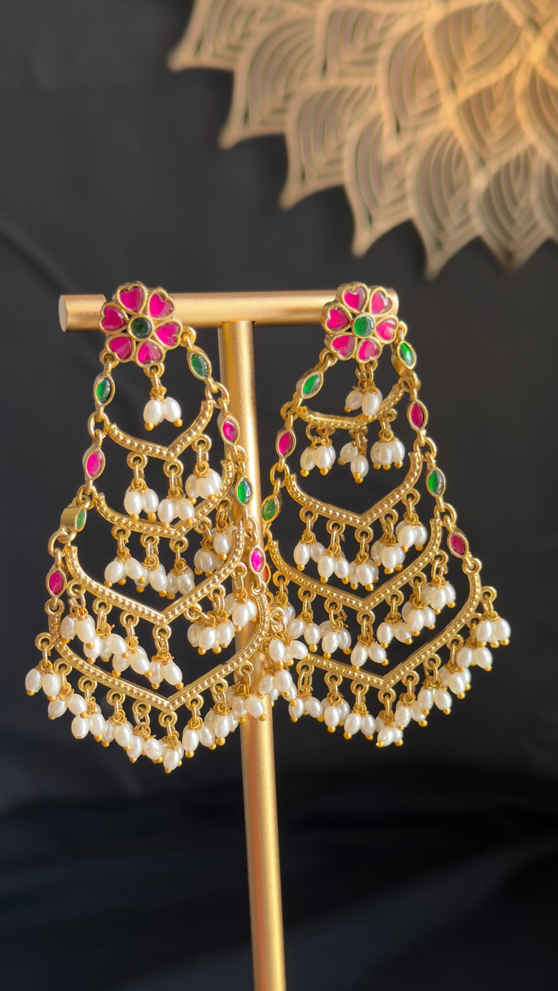 A pair of ornate gold-toned Indian jhumka earrings with white pearl bead drops and pink and green stone accents, displayed on a gold stand.