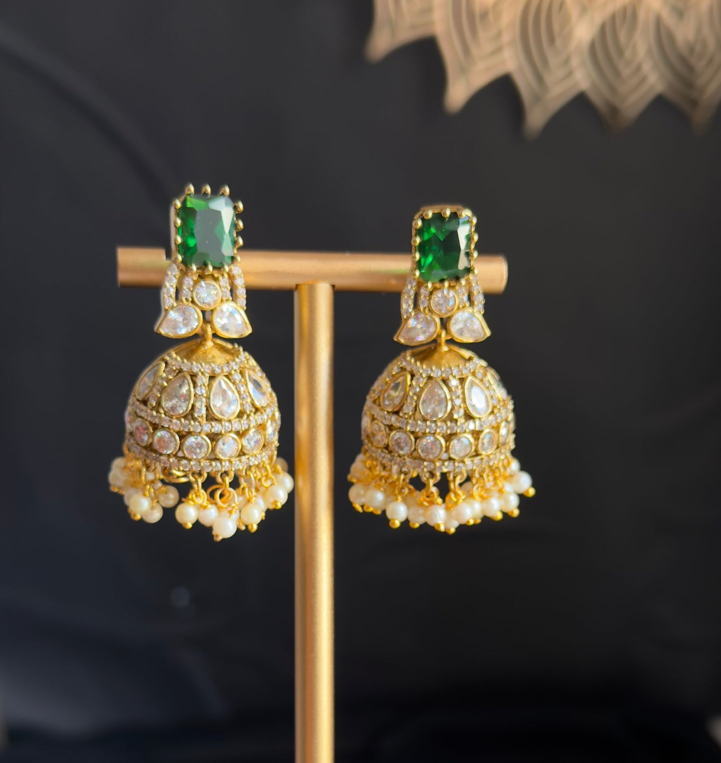 A pair of ornate gold-toned jhumka earrings with rectangular green stones and white stone embellishments, displayed on a gold stand against a dark draped background.