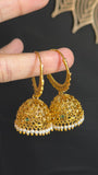 A hand holding a pair of gold-toned traditional Indian jhumka earrings with small green and white stone accents, set against a dark background with a gold mandala design.