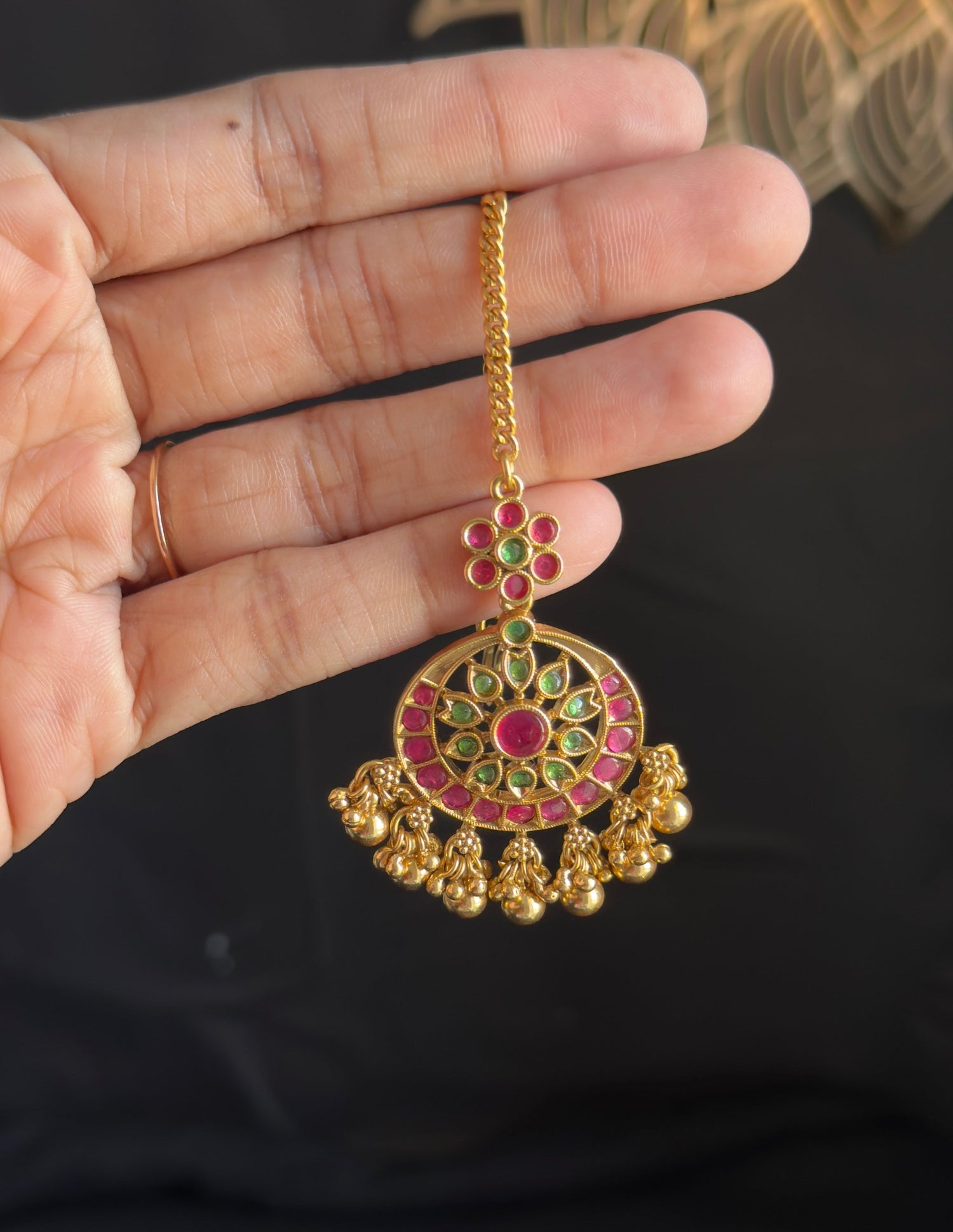 A gold-toned traditional Indian jhumka earring with red and green stone embellishments, held in a person's hand against a dark background.