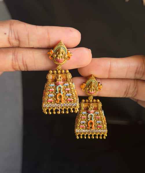 A pair of ornate gold-toned jhumka earrings with colorful enamel detailing, held between two hands against a dark background.