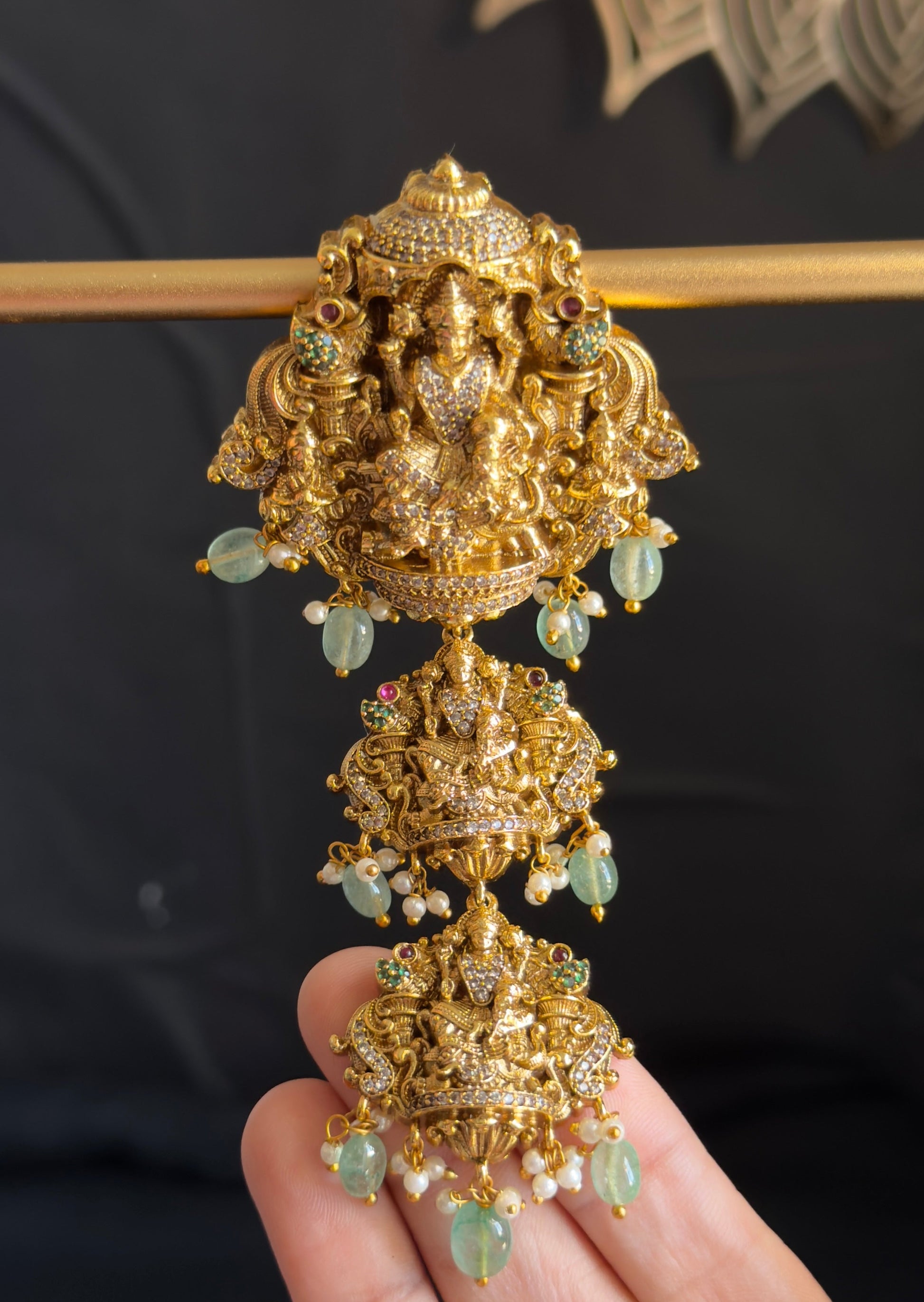 An ornate gold-toned traditional Indian jewelry piece featuring a detailed Ganesha motif and light green stone accents, held in a person's hand against a dark background.