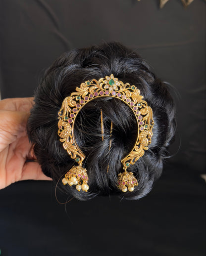 A hand holds a black hair bun decorated with an ornate gold filigree hair clip featuring small green and white stones and dangling jhumka-style ornaments, set against a black background with a gold decorative element in the upper right.