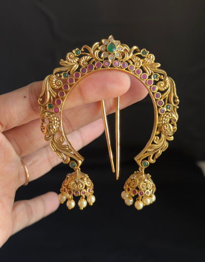 An ornate gold-toned traditional Indian hair ornament (Jhumka headpiece) with dangling Jhumka earrings, held by a hand against a dark background.