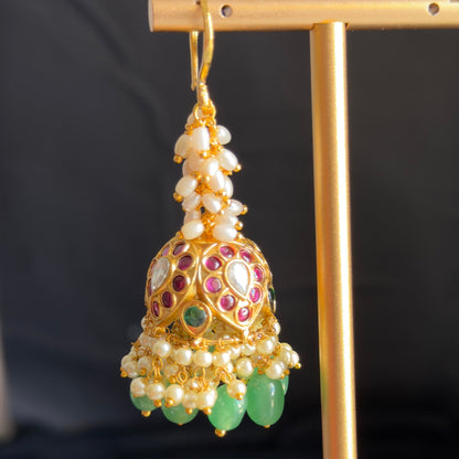 An ornate gold-toned jhumka earring with white pearl-like beads, red and green stone accents, and hanging green teardrop charms, displayed on a gold stand against a dark background.