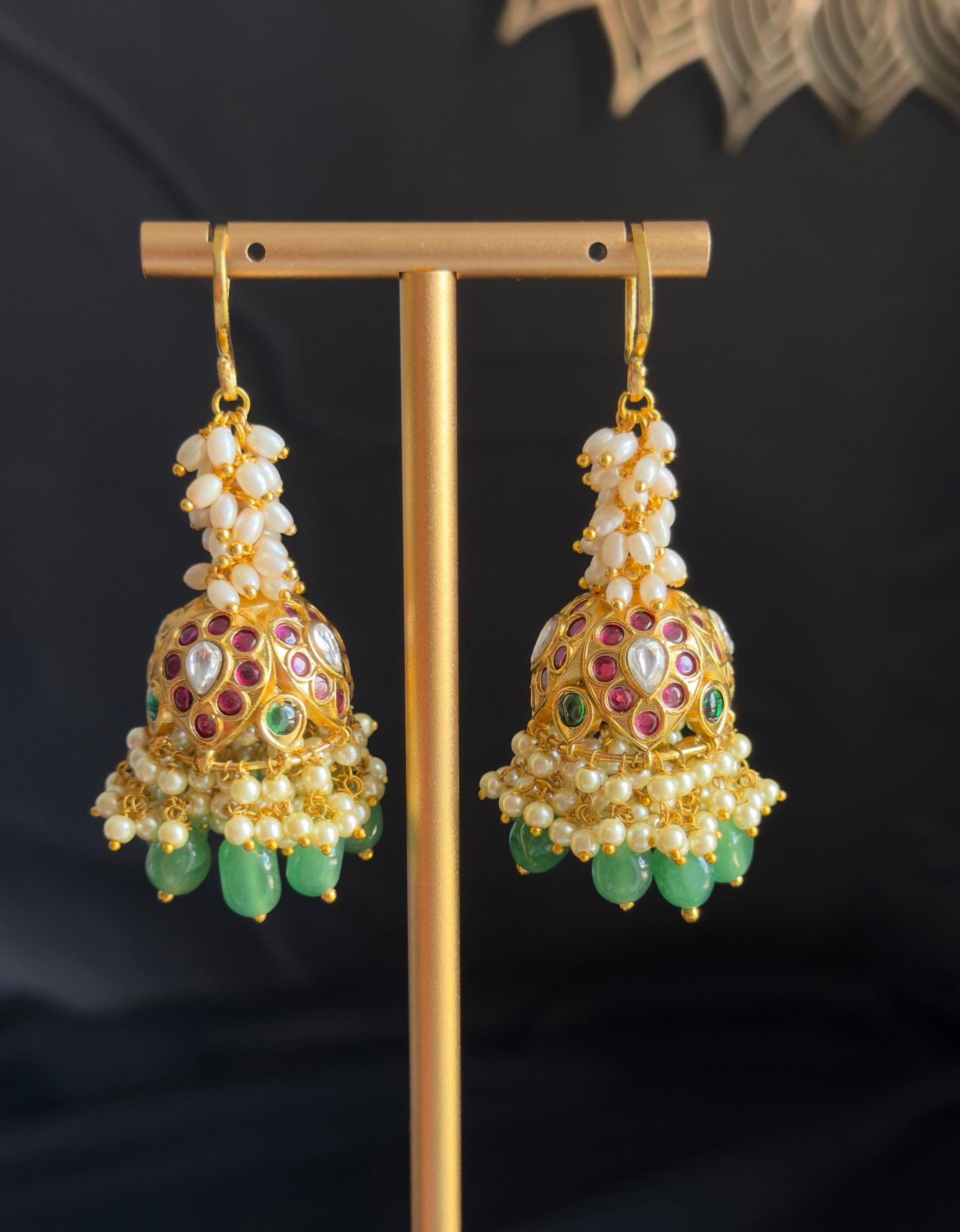 A pair of ornate gold-toned jhumka earrings with white pearl-like beads, multicolored enamel detailing, and dangling green stone accents, displayed on a gold stand against a dark background.
