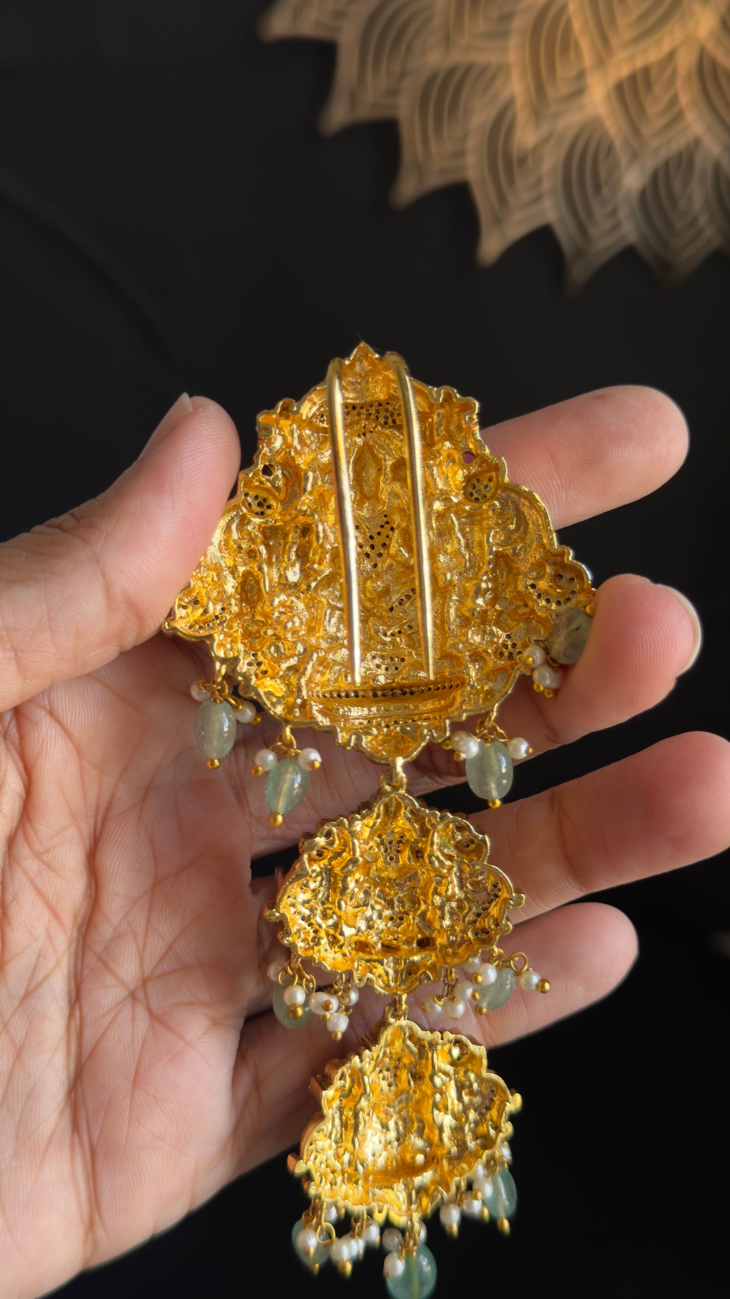 A hand holding a detailed gold-toned traditional pendant brooch with intricate filigree work and hanging green bead accents against a dark background.