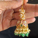 A hand holding a gold-toned traditional Indian jhumka earring decorated with white pearls, green stones, and red and purple enamel details.